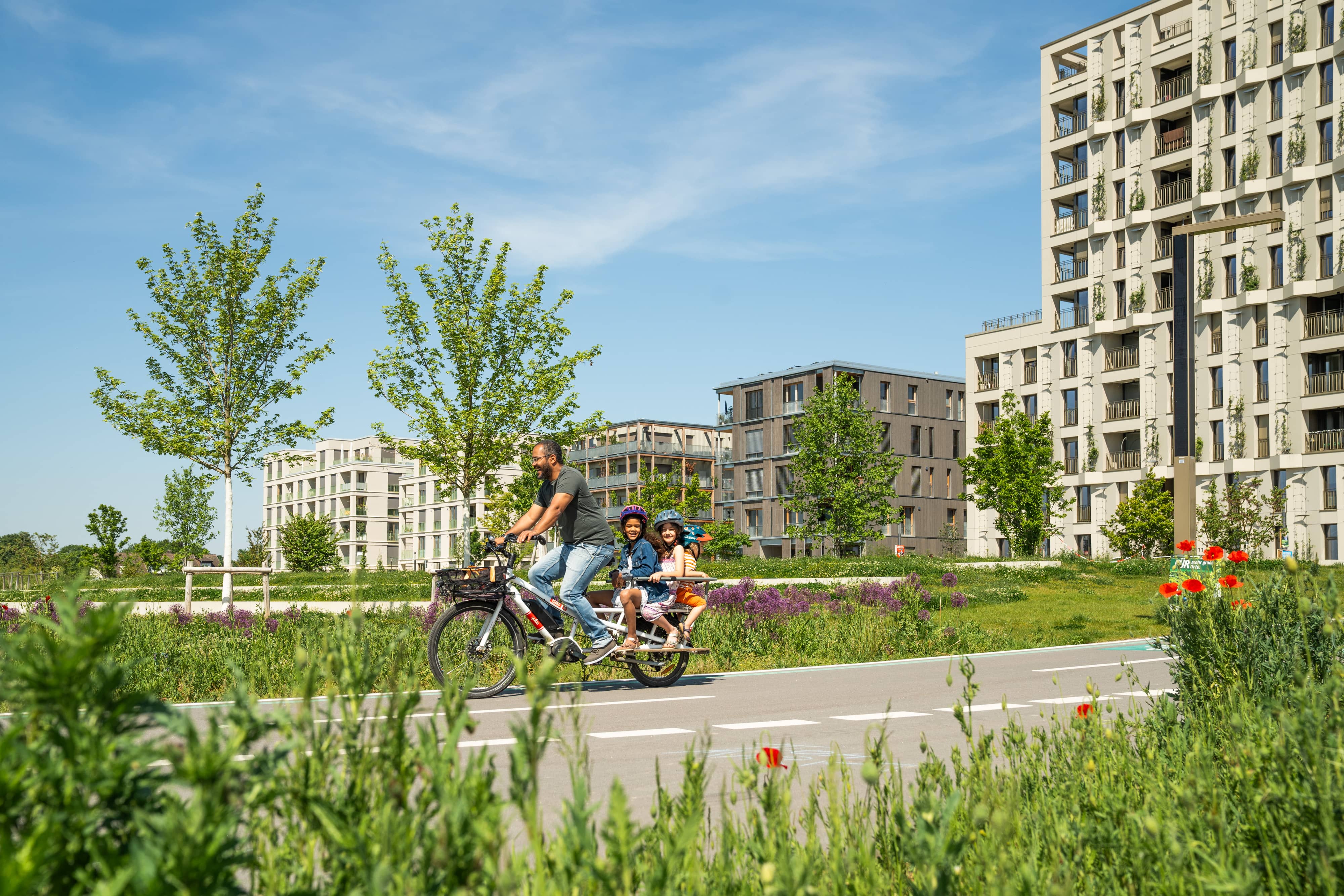 Ein Mann fährt mit drei Kindern auf einem Fahrrad auf dem Radschnellweg durch den Spinelli Park. Im Hintergrund ist die Wohnbebauung zu sehen.