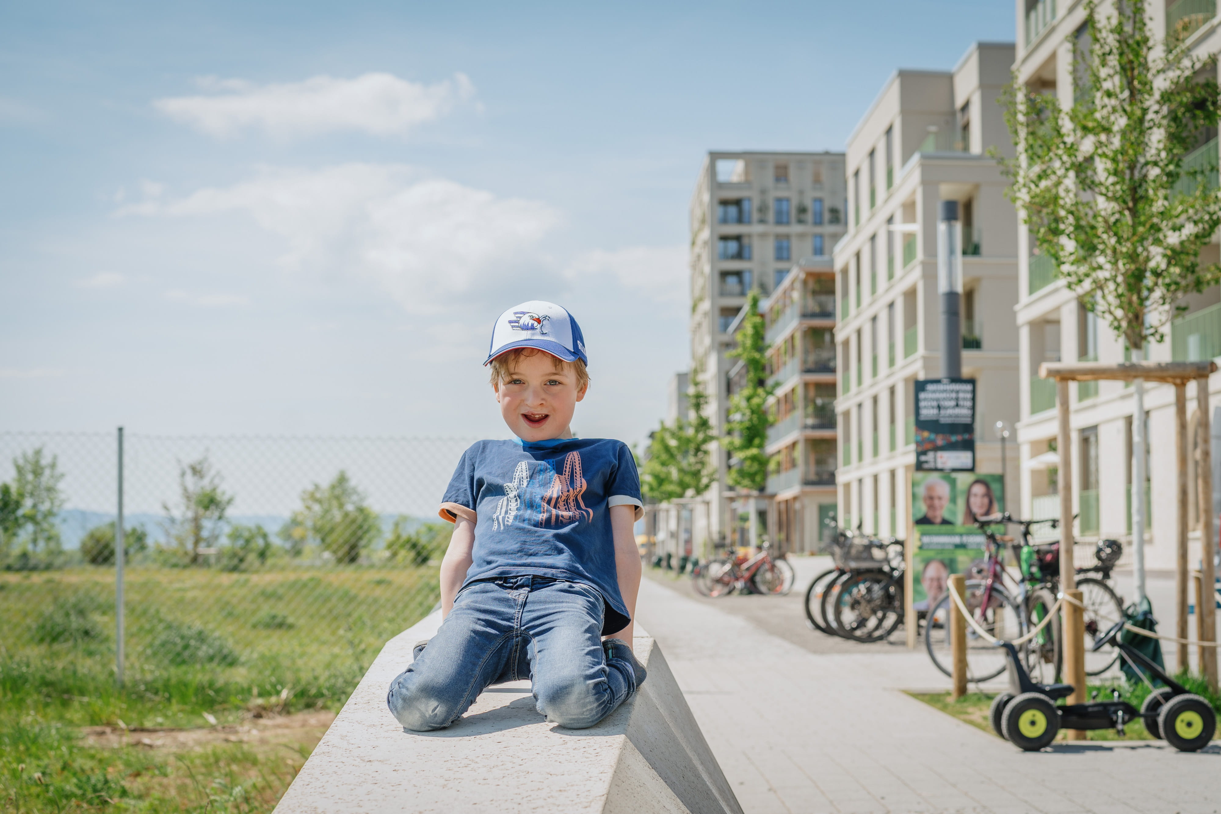 Ein kleiner Junge mit Baseballkappe lächelt in die Kamera. Im Hintergrund ist die Spinelli Wohnbebauung und der Spinelli Park.