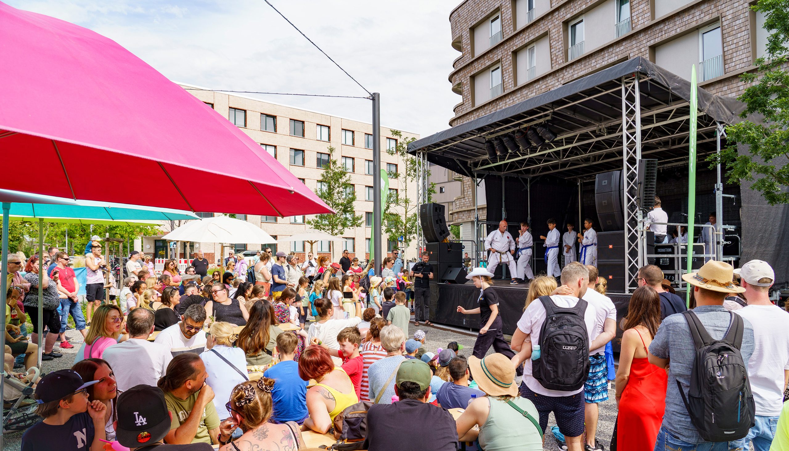 Eine große Menge gemischtes Publikum sitzt an Biertischgarnituren unter bunten Sonnenschirmen und verfolgt eine Bühnendarbietung. Auf einer großen überdachten Bühne befindet sich eine Karategruppe mit vier Kindern und einem erwachsenen Trainer.