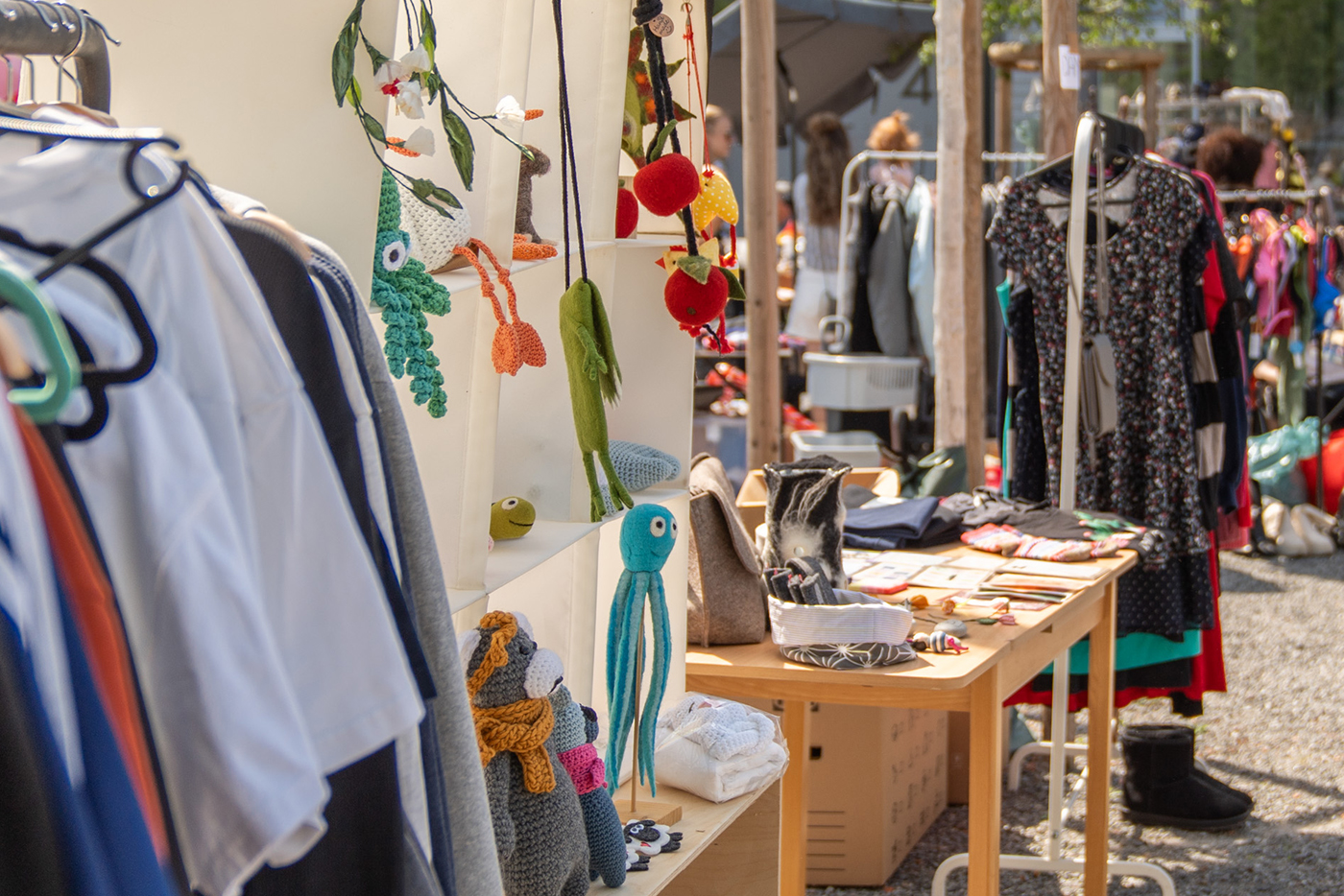 Flohmarktstand mit Holztisch und Regal. Es sind bunte, selbstgehäkelte Kuscheltiere in verschiedenen Tierformen ausgestellt. Auf der linken Seite steht ein Kleiderständer mit auf Bügeln aufgehängten gebrauchten Kleidungsstücken.