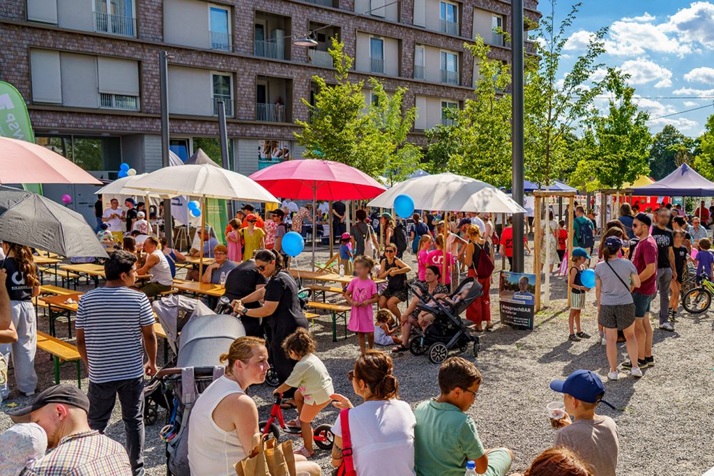 Menschenmenge mit großen Sonnenschirmen und Pavillons auf einem Platz mit jungen Bäumen.