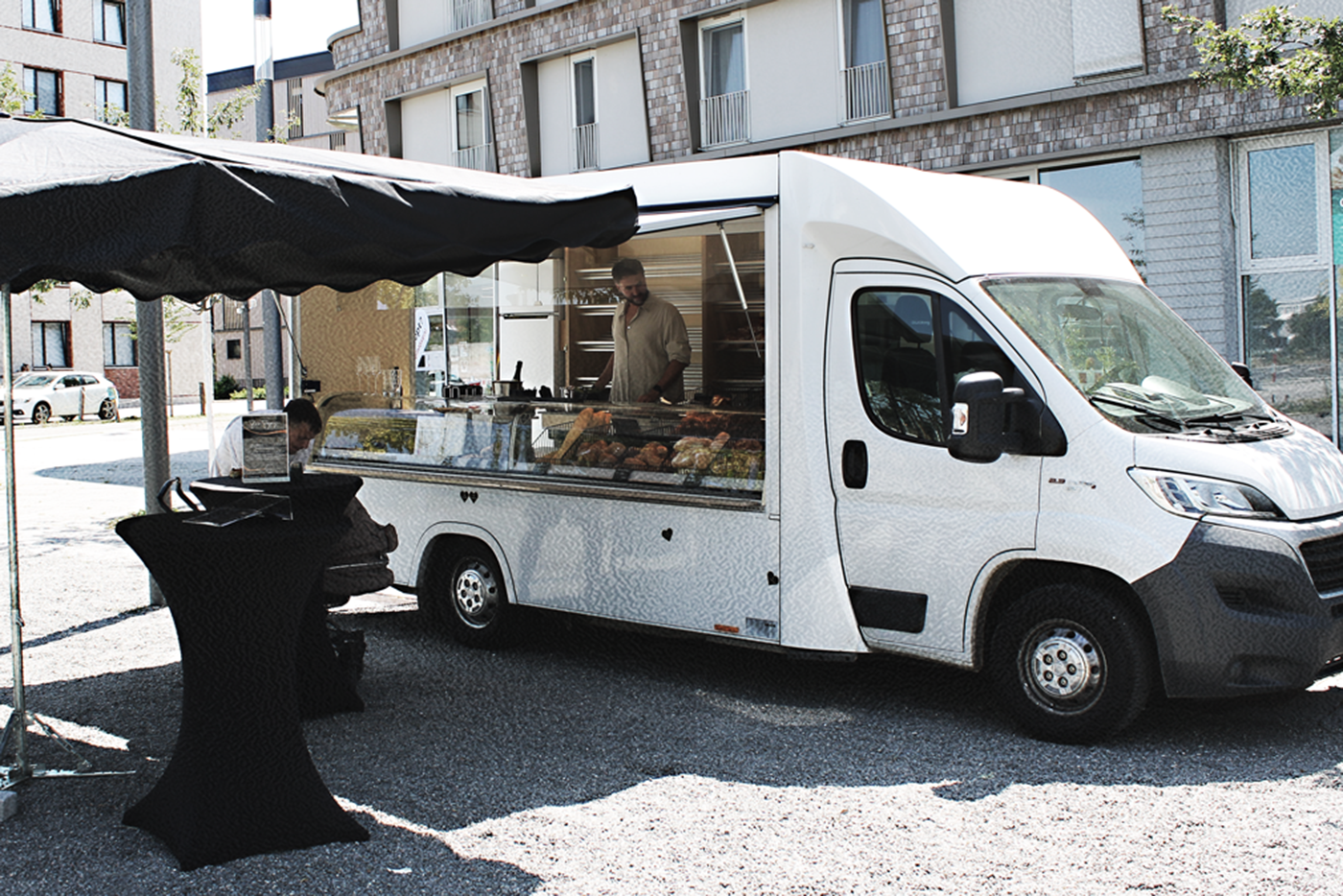 Ein Brotverkaufswagen mit aufgeklapptem Seitenteil parkt vor einem Wohngebäude. Vor dem Wagen befindet sich ein großer Sonnenschirm und ein Stehtisch.