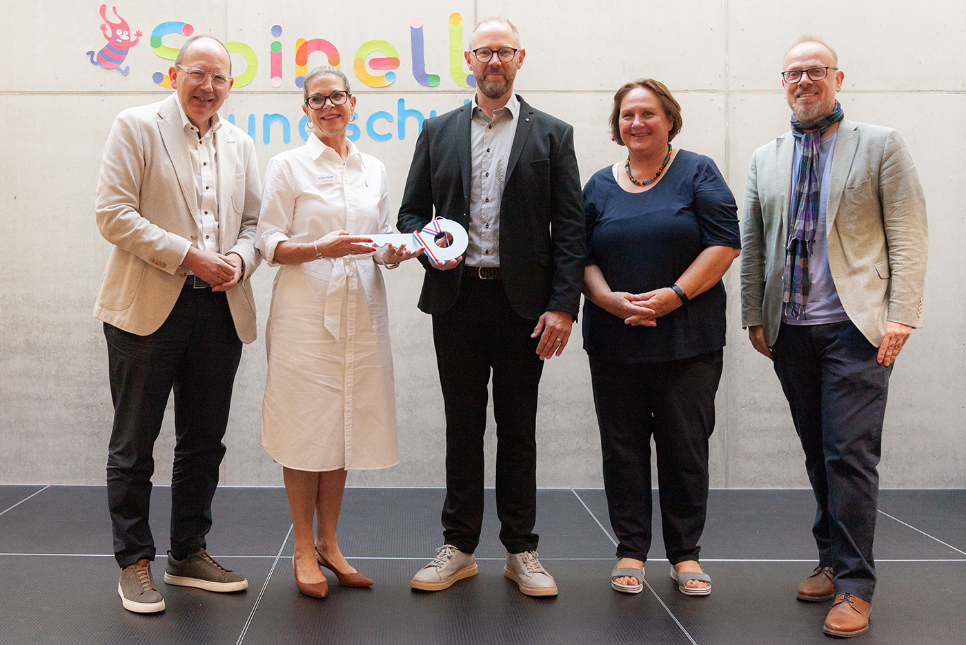 3 Männer und zwei Frauen stehen bei einer symbolischen Schlüsselübergabe nebeneinander vor einer Wand mit der Aufschrift Spinelli Grundschule.