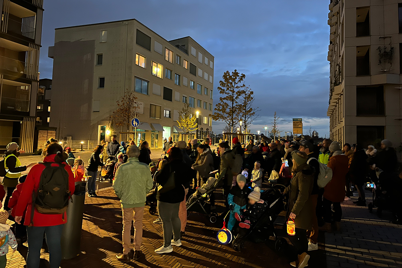 KI-basierte Bildbeschreibung | Eine Menschenmenge, darunter auch Kinder in Kinderwagen, versammelt sich in der Abenddämmerung auf einer Straße in der Nähe von Wohnhäusern, die von Straßenlaternen beleuchtet werden.