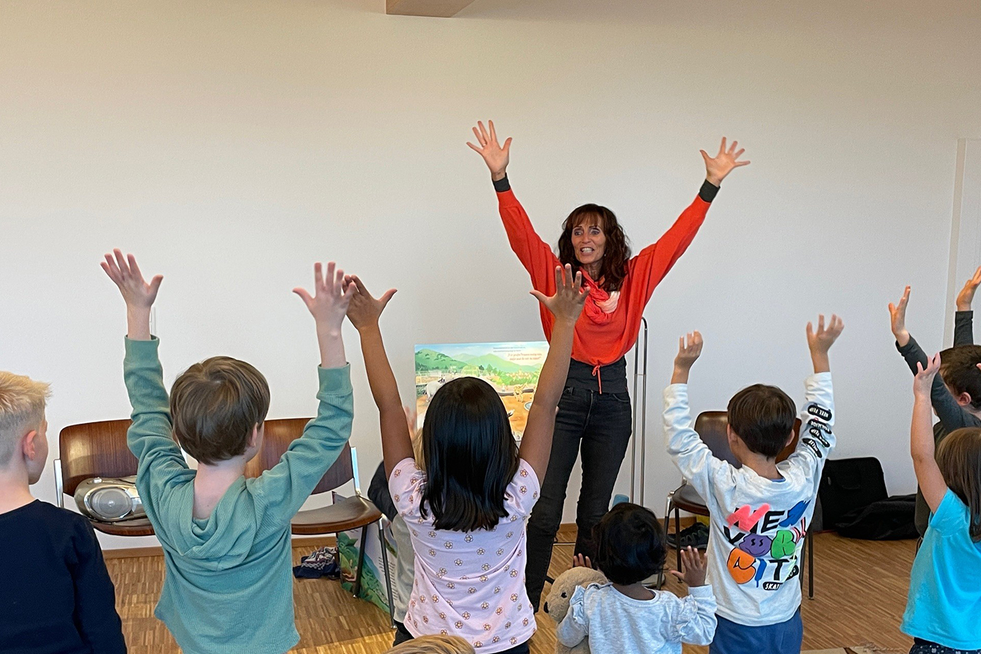 KI-basierte Bildbeschreibung | Eine Frau steht mit erhobenen Armen vor einer Gruppe von Kindern und leitet eine interaktive Aktivität an. Die Kinder ahmen ihre Geste nach und heben ebenfalls ihre Arme.