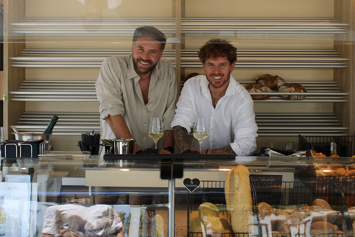 KI-basierte Bildbeschreibung | Zwei Männer stehen hinter einer Bäckertheke mit Brot und Gebäck; beide lächeln, auf der Theke stehen zwei Gläser mit Weißwein.