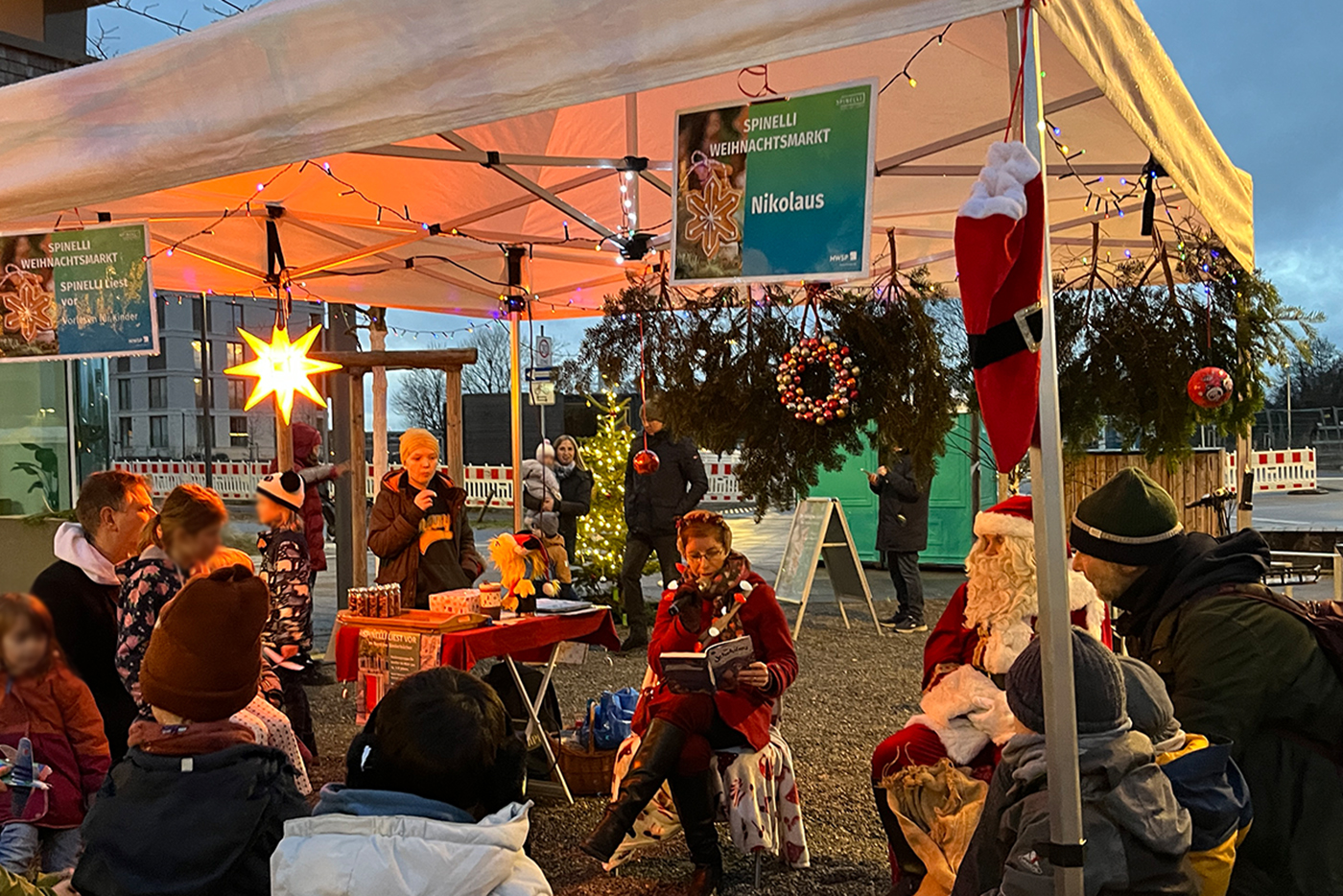 KI-basierte Bildbeschreibung | Eine Gruppe von Kindern und Erwachsenen versammelt sich unter einem mit Weihnachtsschmuck geschmückten Zelt, in dem eine als Weihnachtsmann verkleidete Person sitzt, während eine andere aus einem Buch liest.