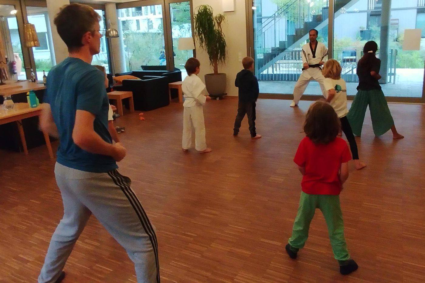 KI-basierte Bildbeschreibung | Eine Gruppe von Kindern und ein Erwachsener üben in einem hellen Raum mit Holzfußboden und großen Fenstern Kampfsportarten.