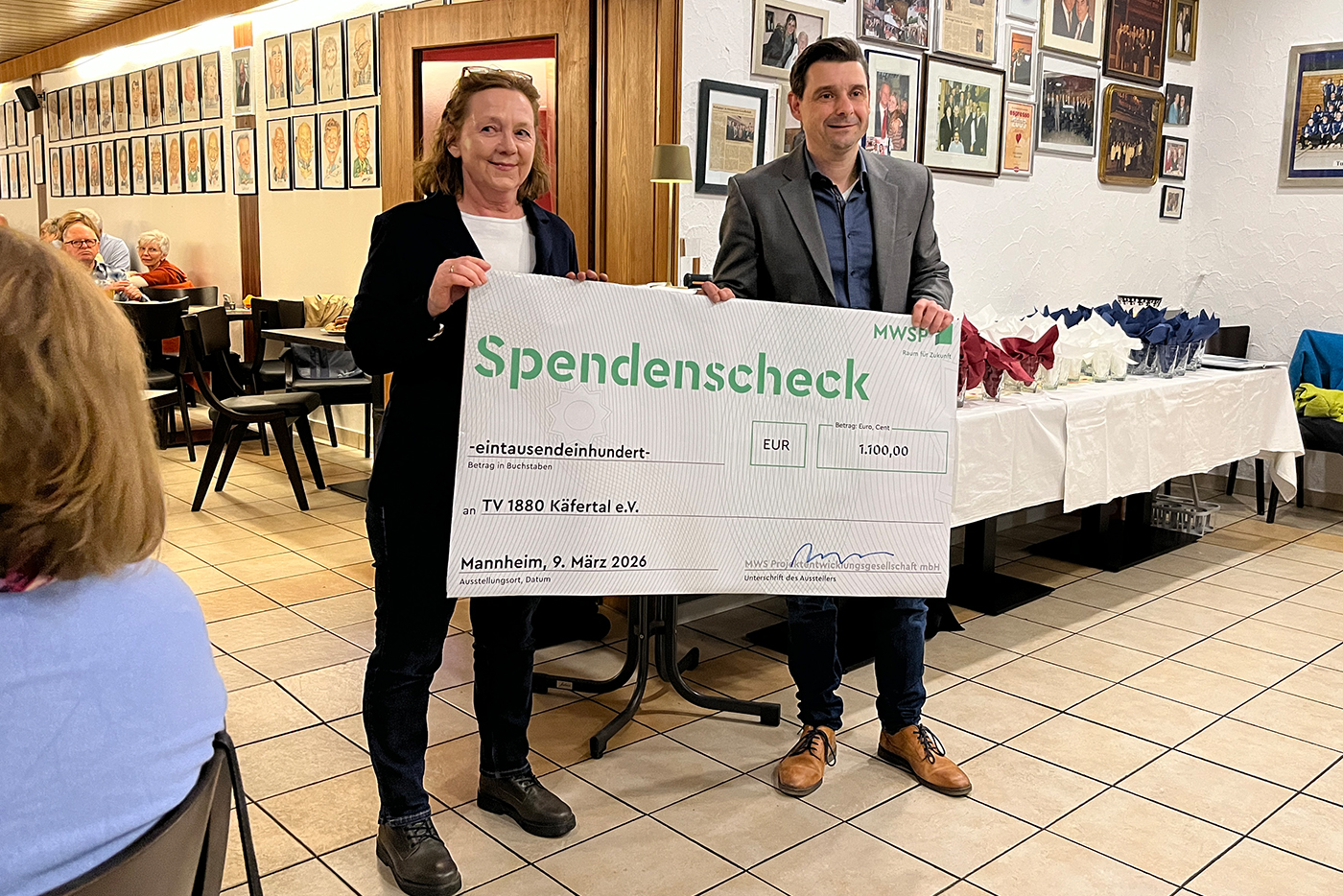 KI-basierte Bildbeschreibung | Zwei Personen stehen drinnen und halten einen großen feierlichen Scheck über 1.100 Euro mit der Aufschrift "Spendenscheck" in der Hand, während andere an Tischen im Hintergrund sitzen.