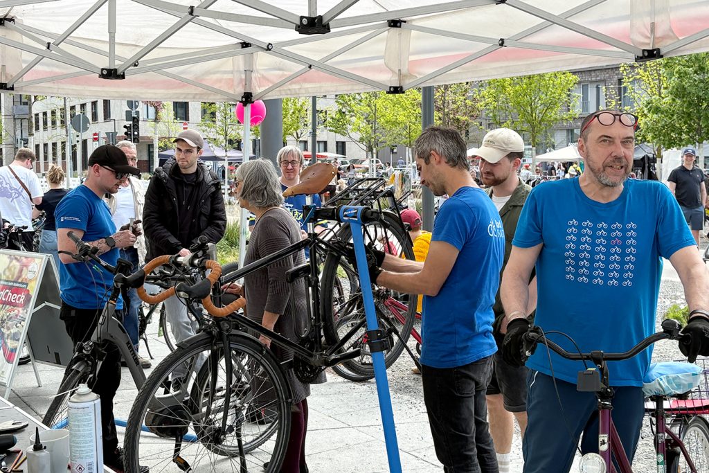 KI-basierte Bildbeschreibung | Mehrere Personen versammeln sich unter einem weißen Zeltdach bei einer Veranstaltung im Freien, wo Mechaniker Fahrräder an Ständern reparieren.