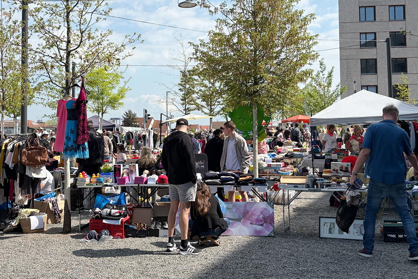 KI-basierte Bildbeschreibung | Flohmarkt im Freien, bei dem die Besucher bei sonnigem Wetter auf Tischen verschiedene Artikel wie Kleidung, Haushaltswaren und Dekorationen stöbern.