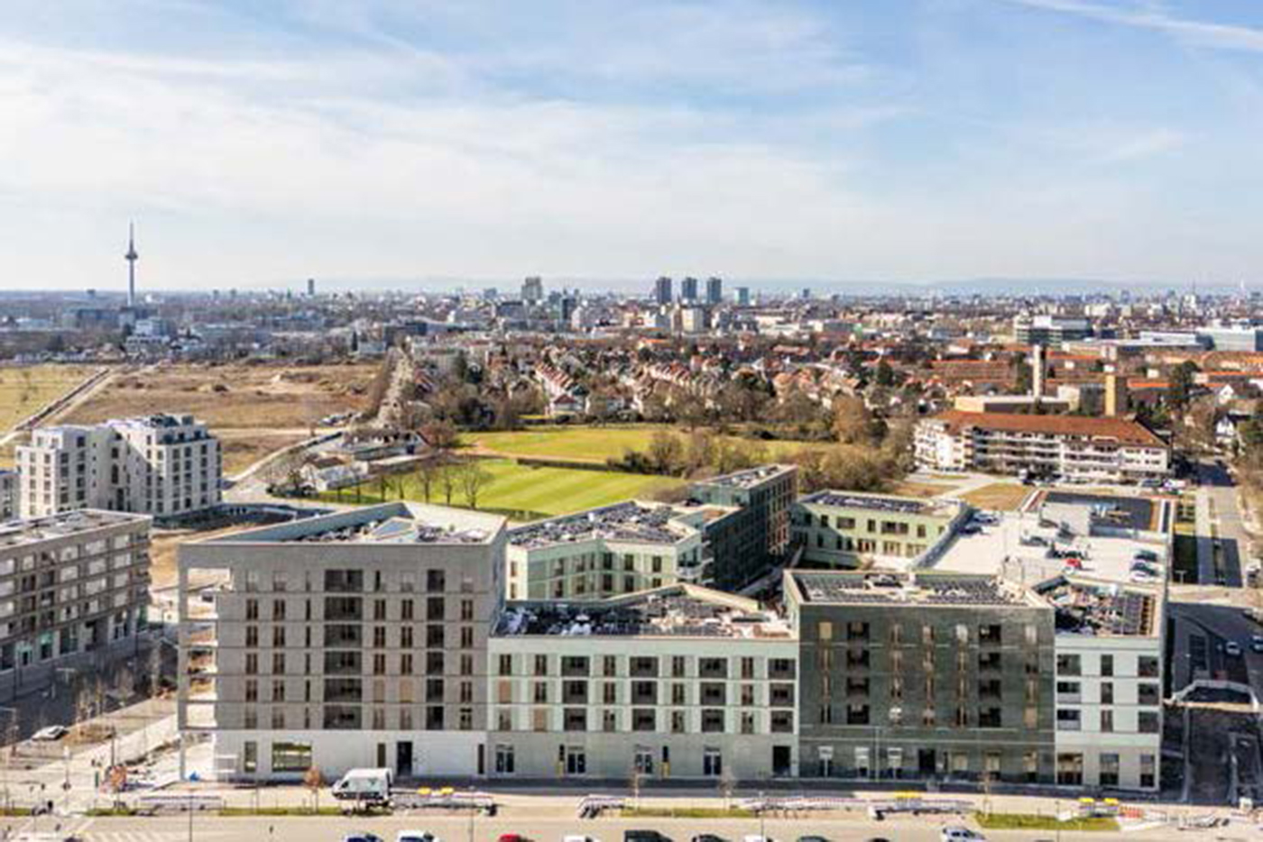 KI-basierte Bildbeschreibung | Luftaufnahme eines modernen städtischen Wohnkomplexes mit Grünflächen, umgeben von Straßen und städtischen Gebäuden, unter einem teilweise bewölkten Himmel - der Bau schreitet in Richtung der kommenden Termine stetig voran.