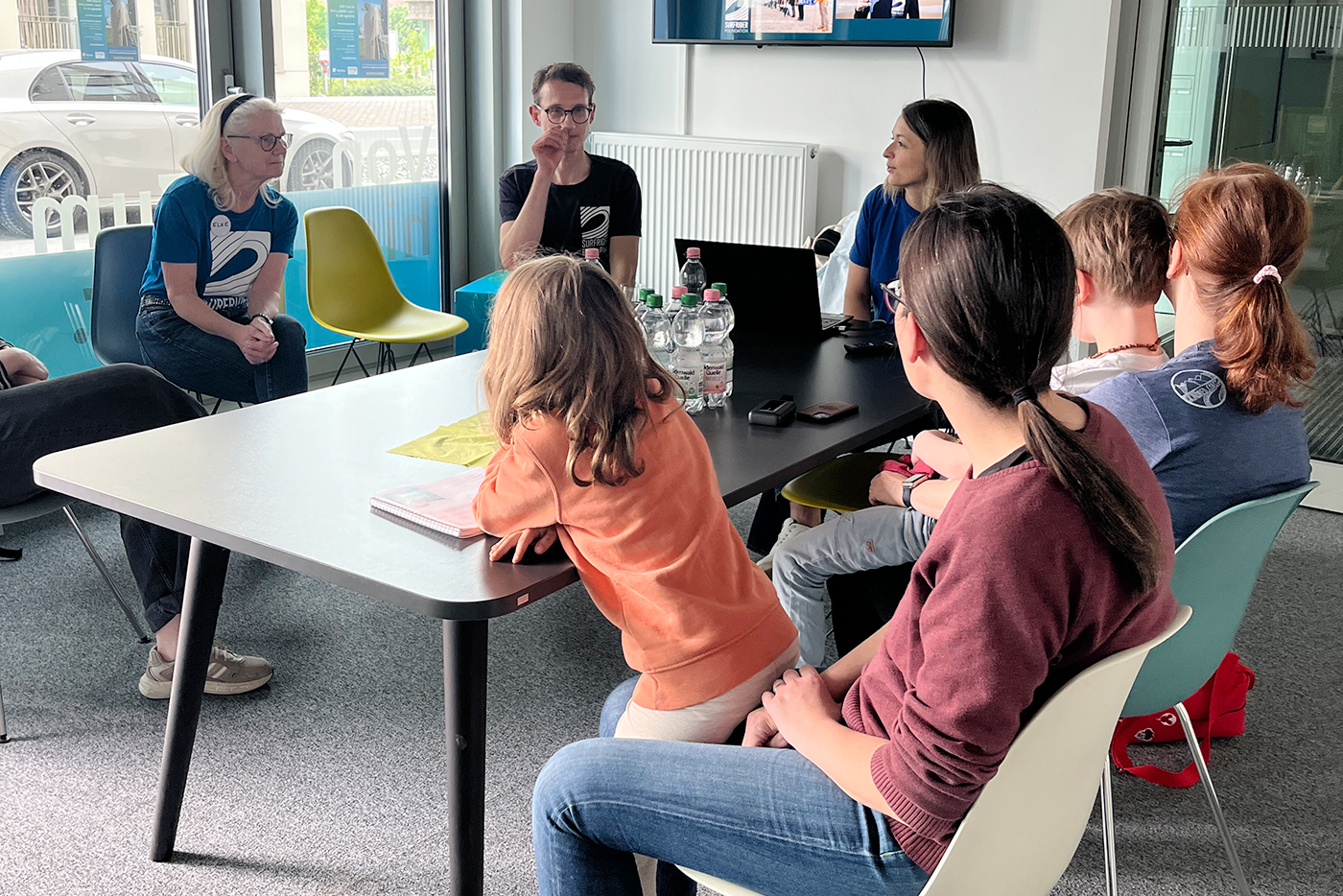 KI-basierte Bildbeschreibung | Eine Gruppe von Personen sitzt in einem Besprechungsraum um einen Tisch herum und führt ein Gespräch. Auf dem Tisch stehen Wasserflaschen und Papiere.