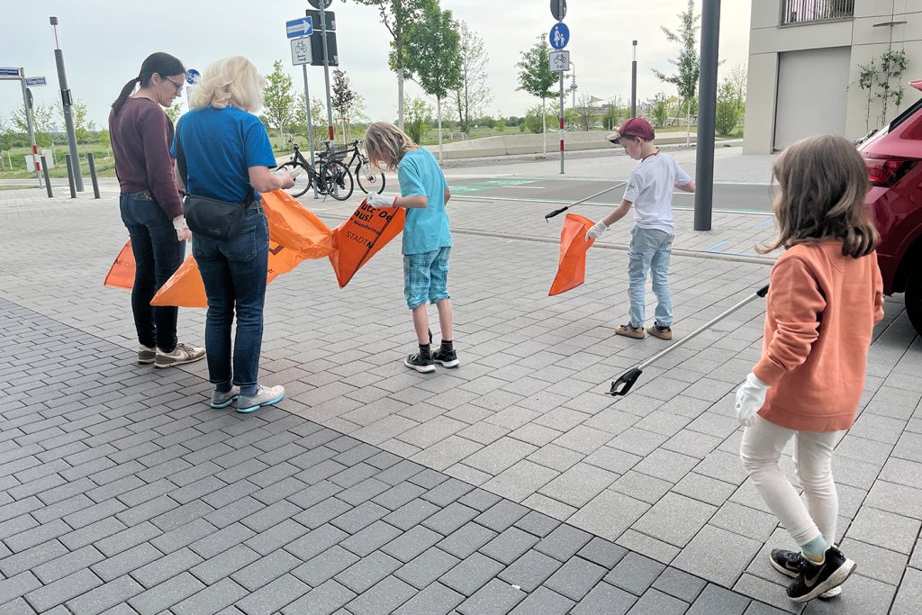 KI-basierte Bildbeschreibung | Eine Gruppe von Erwachsenen und Kindern sammelt mit Greifern und orangefarbenen Säcken Müll auf einem gepflasterten Gehweg in der Nähe von geparkten Autos und Fahrrädern auf.