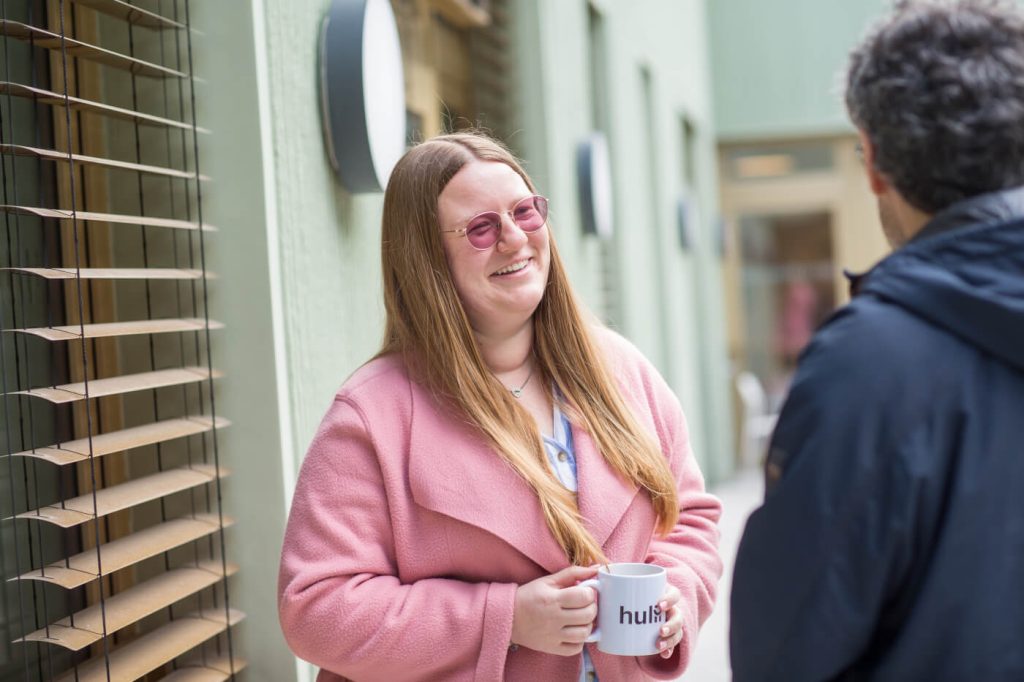 KI-basierte Bildbeschreibung | Eine Frau mit einer rosafarbenen Brille und einem rosafarbenen Mantel lächelt, während sie eine Tasse in der Hand hält und mit einer Person im Freien spricht.