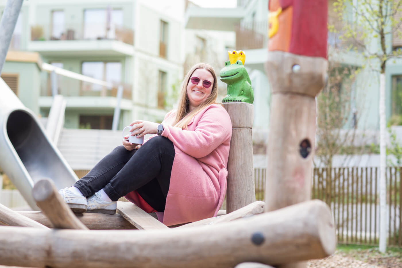 KI-basierte Bildbeschreibung | Eine Frau in einem rosafarbenen Mantel und mit Sonnenbrille sitzt auf einem hölzernen Spielplatzgerüst und hält einen Becher neben einer grünen Froschstatue, die eine Krone trägt.
