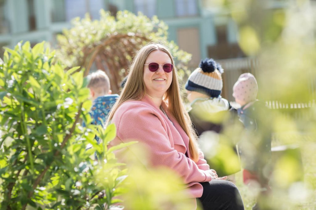 KI-basierte Bildbeschreibung | Eine Frau in einem rosa Mantel und mit einer runden Sonnenbrille sitzt lächelnd im Freien, während im Hintergrund Kinder an einem sonnigen Tag spielen.
