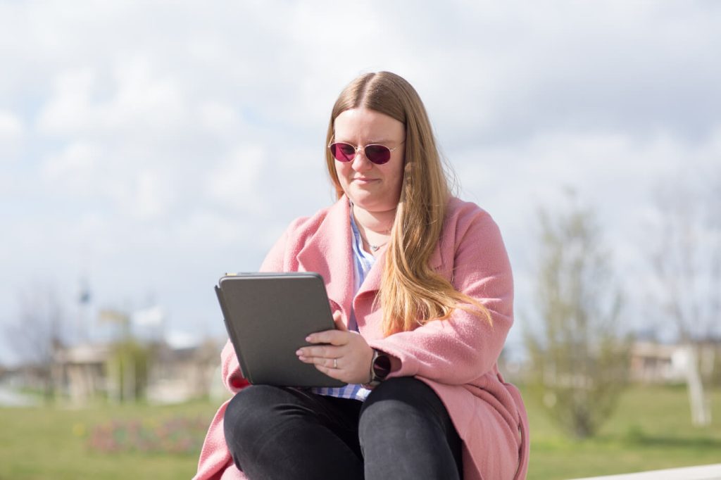 KI-basierte Bildbeschreibung | Eine Frau mit rosa Sonnenbrille und rosa Mantel sitzt im Freien und benutzt ein Tablet.