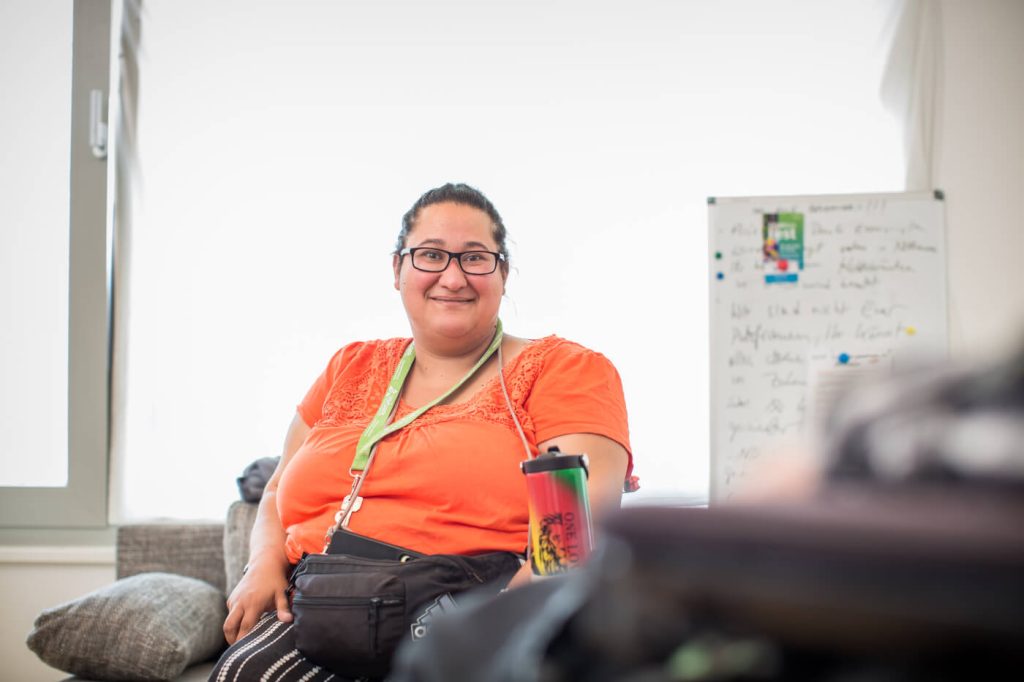 KI-basierte Bildbeschreibung | Eine Person mit Brille und orangefarbenem Hemd sitzt im Haus und lächelt, während im Hintergrund ein Reisebecher und ein Whiteboard zu sehen sind.