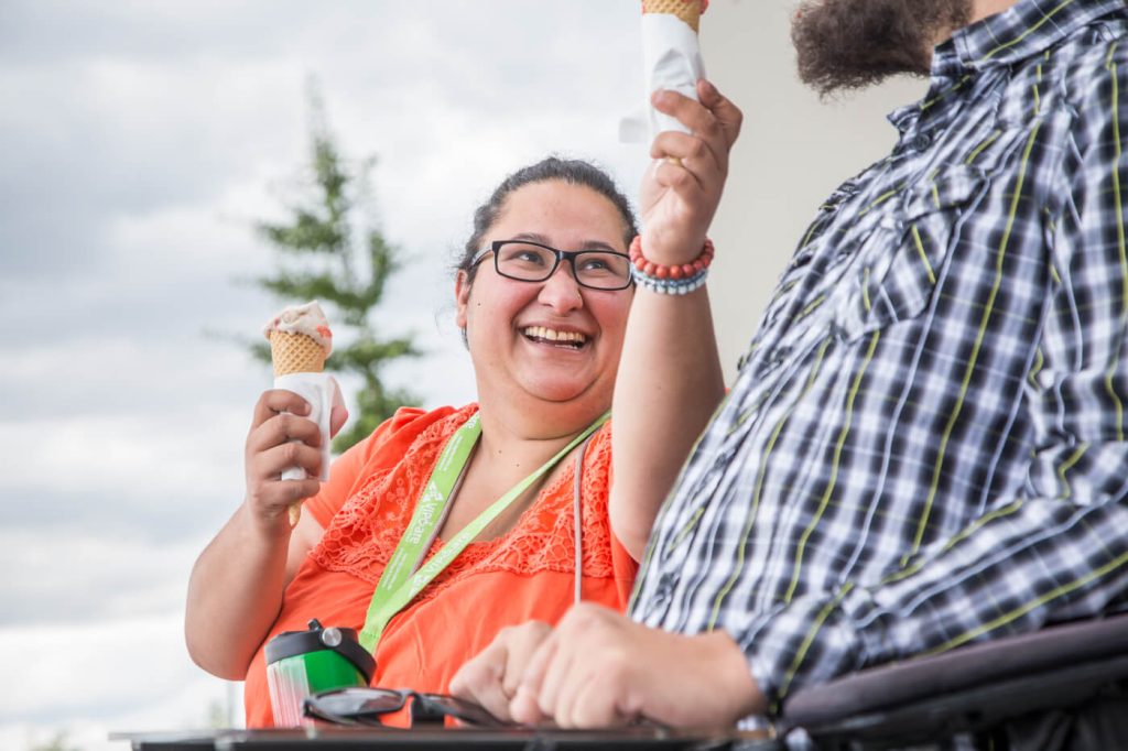 KI-basierte Bildbeschreibung | Zwei Personen sitzen im Freien, lächeln und halten Eiswaffeln in der Hand. Eine Person trägt ein orangefarbenes Hemd und eine Brille, während sie die andere Person, die ein kariertes Hemd trägt, anschaut.