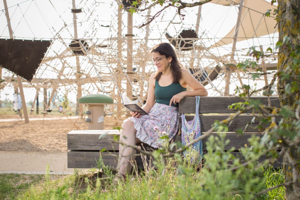 KI-basierte Bildbeschreibung | Eine Frau sitzt auf einer Bank und liest ein Buch in einem Park in der Nähe eines Spielplatzes, mit einer bunten Tragetasche an ihrer Seite.