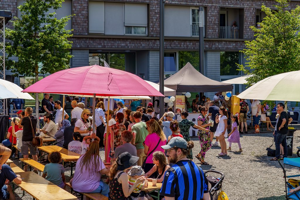 KI-basierte Bildbeschreibung | An einem sonnigen Tag versammeln sich Menschen im Freien bei einer Gemeinschaftsveranstaltung mit Zelten, Sonnenschirmen, Bänken und Kindern vor dem Hintergrund eines Wohnhauses.