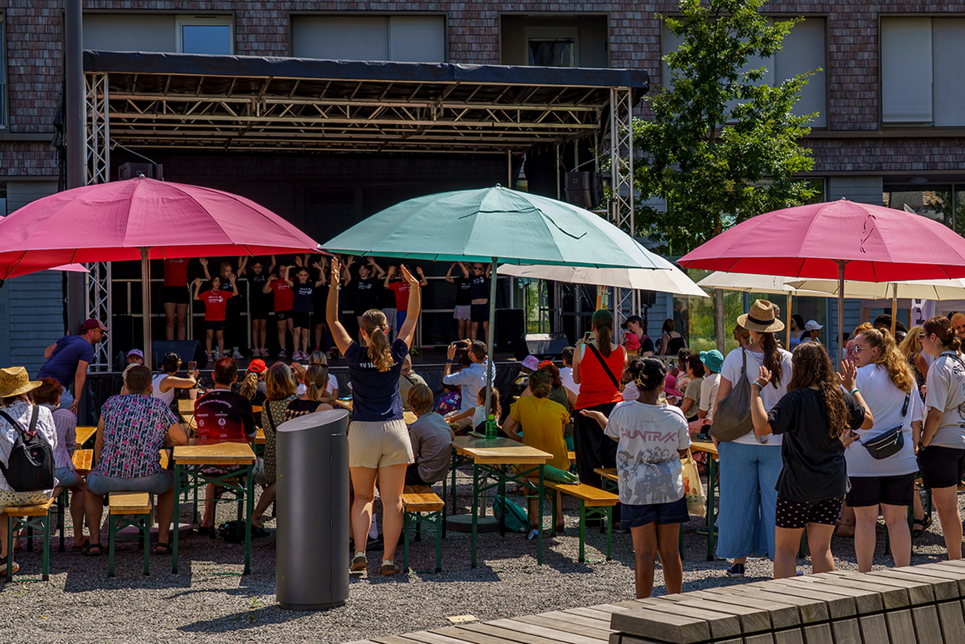KI-basierte Bildbeschreibung | An einem sonnigen Tag sitzen die Zuschauer an Picknicktischen unter großen Sonnenschirmen und verfolgen eine Gruppe von Künstlern auf einer Freiluftbühne.
