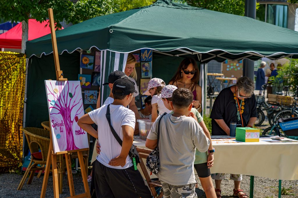 KI-basierte Bildbeschreibung | Kinder und Erwachsene unterhalten sich an einem Kunststand im Freien unter einem grünen Vordach, an dem Gemälde ausgestellt sind und auf dem Malutensilien bereitliegen.