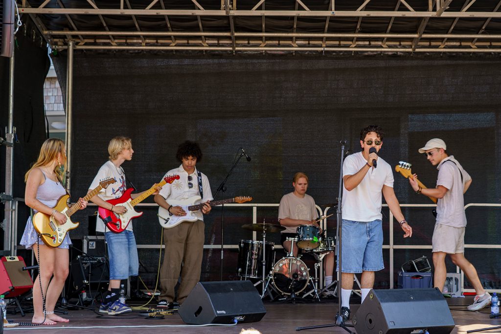 KI-basierte Bildbeschreibung | Eine fünfköpfige Band tritt auf einer Freiluftbühne auf – mit Schlagzeug, E-Gitarren und einem Sänger, der ein Mikrofon in der Hand hält.